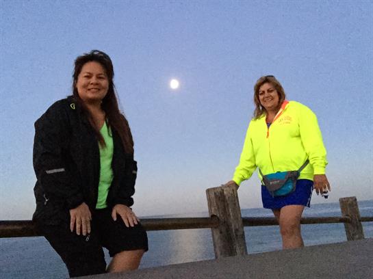 Two women pose on a pier at dusk, enjoying the peaceful waterfront and the glowing moon.