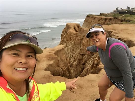 Two smiling hikers point at the rock formations and ocean view while enjoying a cloudy day outdoors.