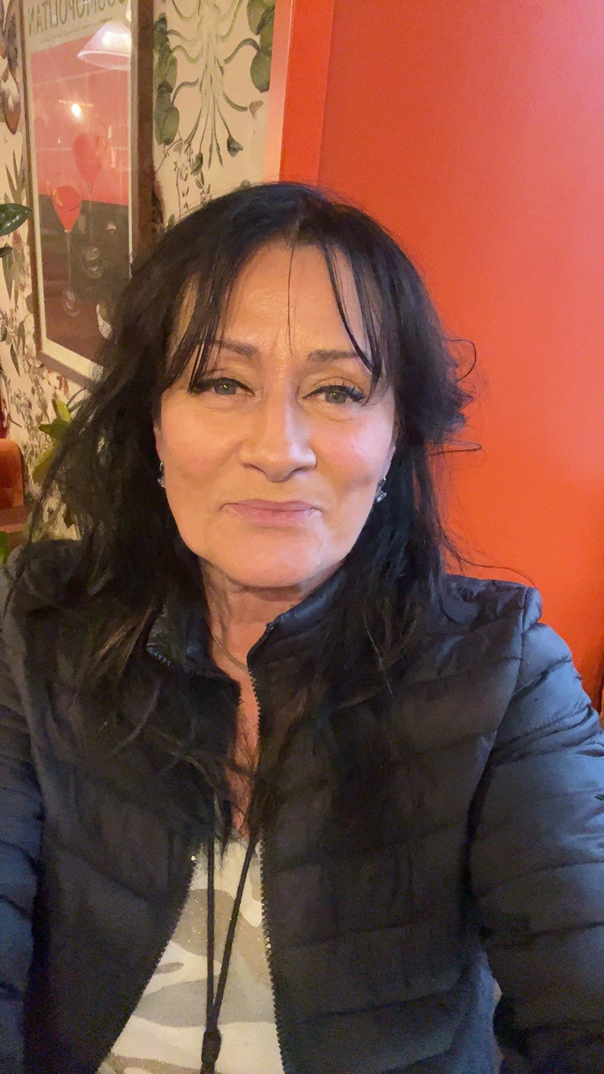 A woman with long dark hair smiles warmly while seated in a colorful indoor space.