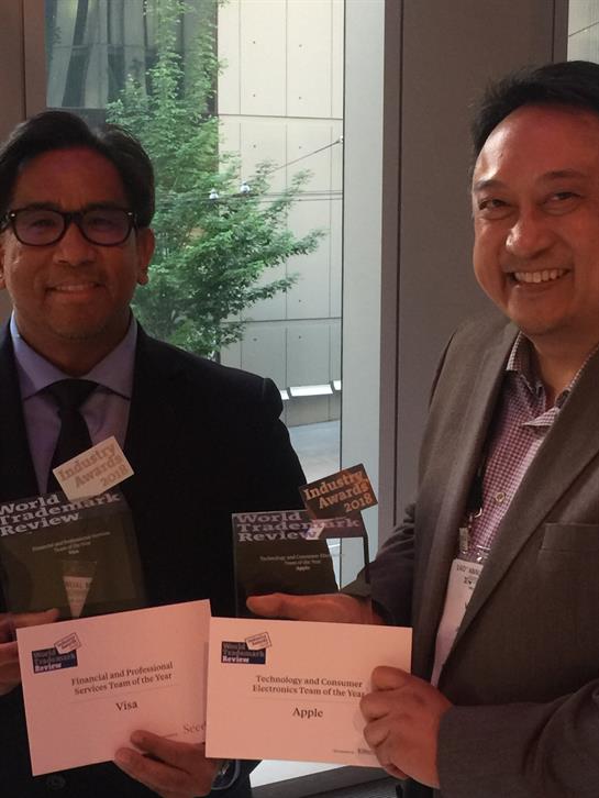 Two professionals pose for a photo, displaying their awards at a conference location with greenery.