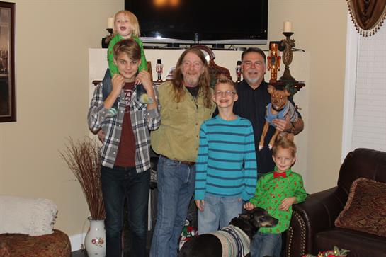 A family of five smiles, kids on shoulders and pets at their feet, creating a joyful scene.