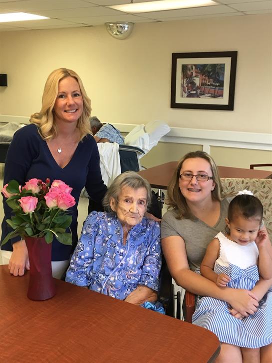 Three generations of women gather around a table in a nursing home, smiling and sharing joy.