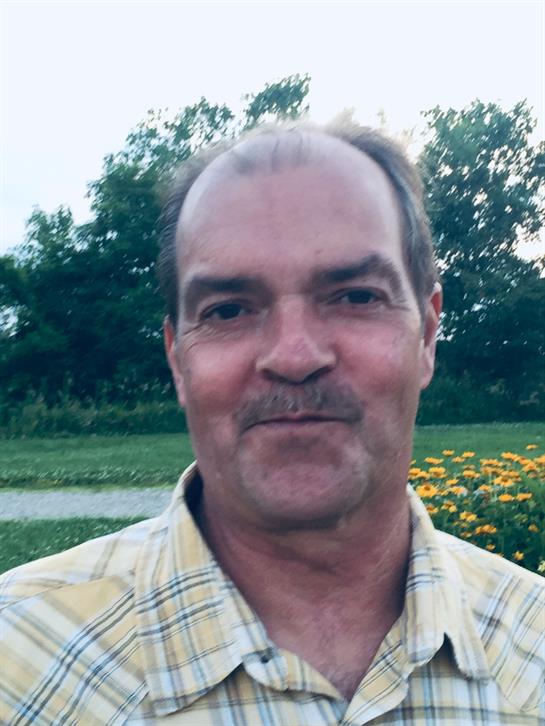 A man stands smiling in a lush park filled with flowers and trees during summer.