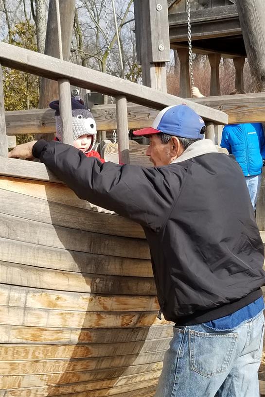 A man engages playfully with a child at a wooden boat play structure in a park setting.