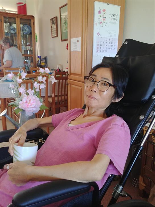 A woman with glasses sits comfortably in a wheelchair, sipping tea and surrounded by flowers.