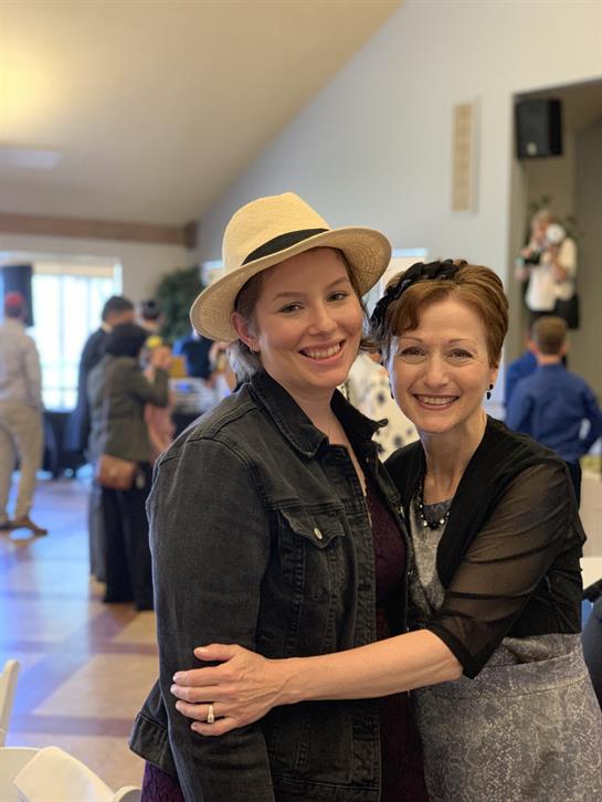 Two women embrace and smile while surrounded by guests at a lively social event indoors.