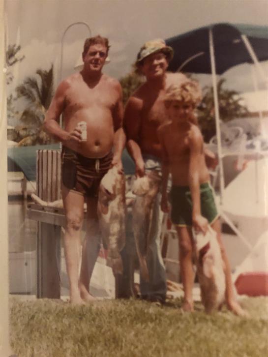 Three men and a boy stand proudly with their catch of fish by the water in tropical surroundings.