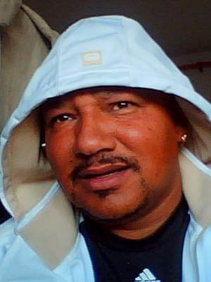 A man with a friendly expression is seated indoors, wearing a hooded jacket and looking content.