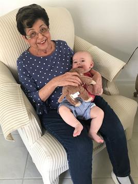 An elderly woman smiles while holding a baby on her lap in a cozy living room setting.