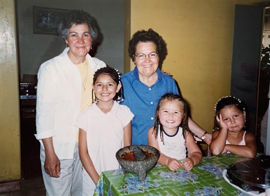 Two grandmothers and three girls are smiling together at a table in a cozy home.