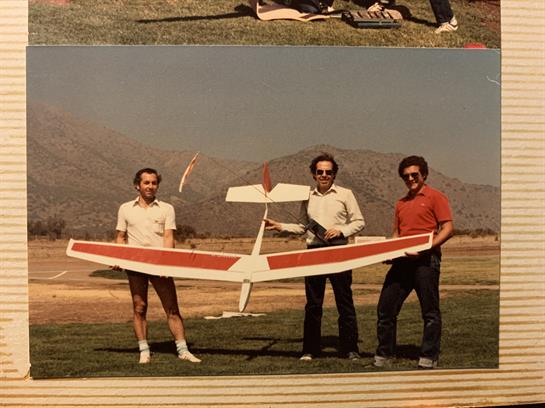 Three men hold a large glider under a clear sky with mountains in the background.