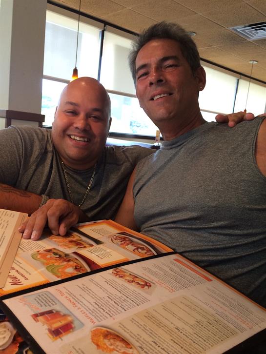 Two men are smiling broadly while sharing a meal and looking at menus in a restaurant.
