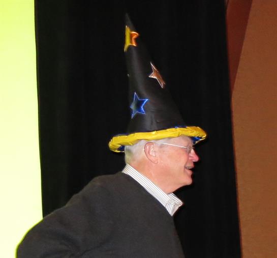 An elderly man enjoys a lively event while wearing a colorful wizard hat adorned with stars.