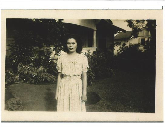 A woman stands in a lush garden, dressed in a vintage dress, surrounded by greenery and sunlight.
