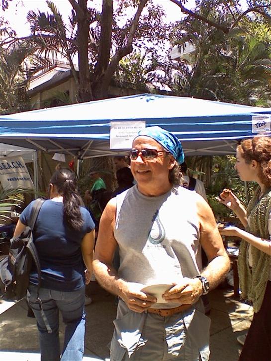 A cheerful man in a gray tank top and blue bandana interacts with visitors at a vibrant market.