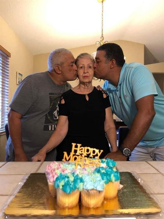 Two men kiss a woman on both cheeks, celebrating Mother's Day with a colorful cake on the table.