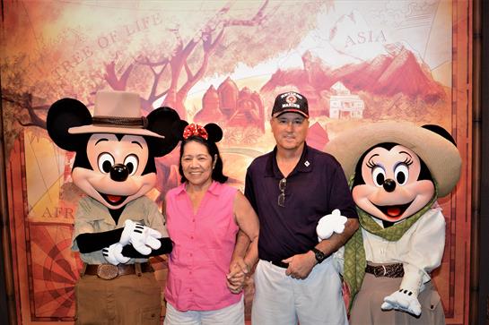 A man and woman smile while posing with Mickey Mouse and Minnie Mouse at a park location.