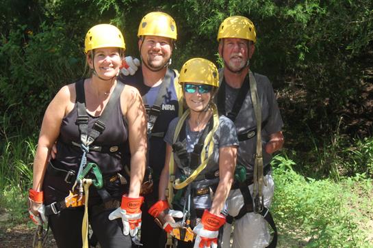 Four friends smile as they prepare for an exciting ziplining adventure in a lush forest.