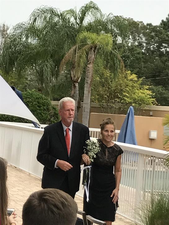 A man and woman stroll together through a lush garden, dressed elegantly for a special occasion.