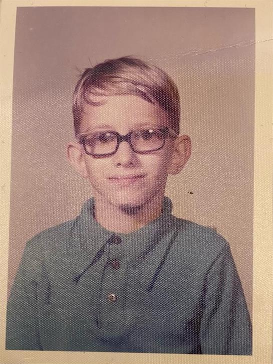A young boy with blonde hair and glasses smiles at the camera, radiating innocence.