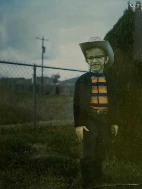 A cheerful young cowboy poses near a fence in a rural setting, displaying a lively spirit.