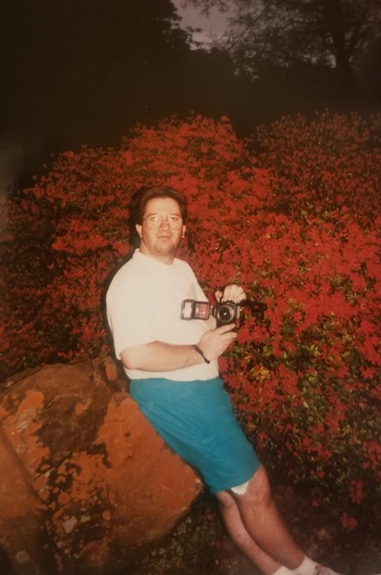 A man stands against a backdrop of bright red bougainvillea flowers while holding a camera.