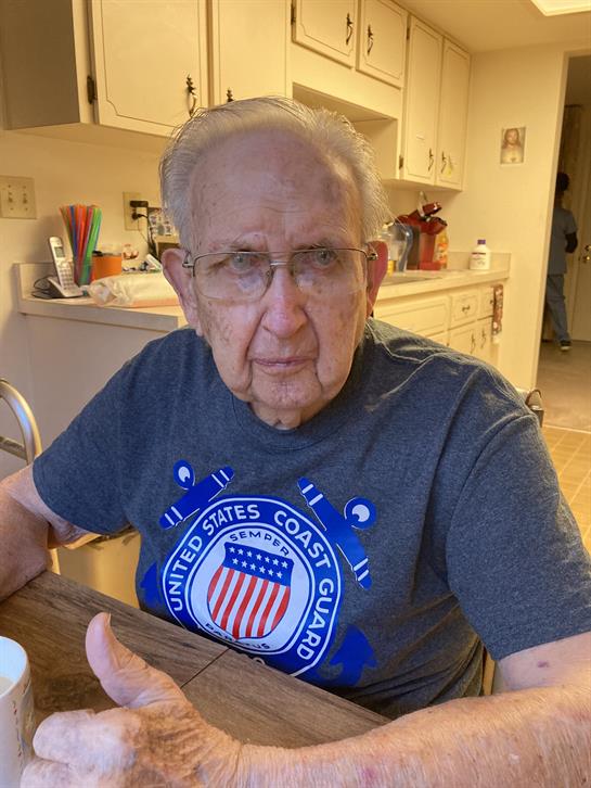 Older gentleman wears a Coast Guard shirt, engaging in conversation at a kitchen table.