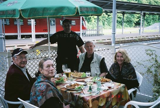 Friends sit together at a colorful table enjoying food and drinks under an umbrella.
