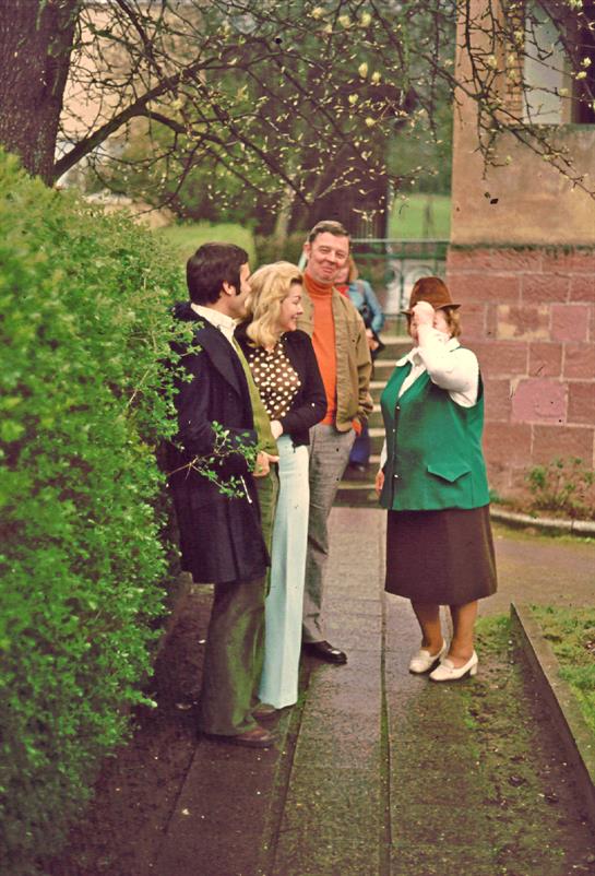 Four individuals are engaged in conversation next to a vibrant hedge while enjoying the outdoors.