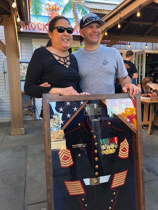 Two people proudly show a framed military uniform at a lively outdoor social event.