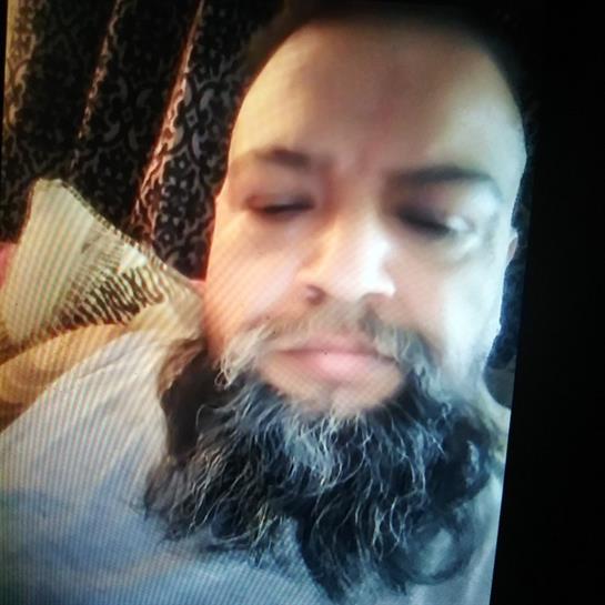A man with a thick beard sits comfortably on a couch surrounded by soft pillows.