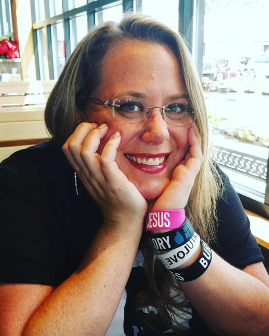 A woman with glasses sits in a cafe, smiling cheerfully while resting her head on her hands.