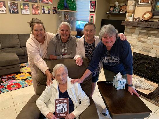 Five family members celebrate a special occasion with their grandmother in a warm living room.