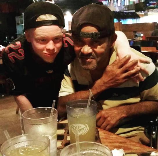 A boy smiles while hugging an older man at a busy restaurant, enjoying drinks together.