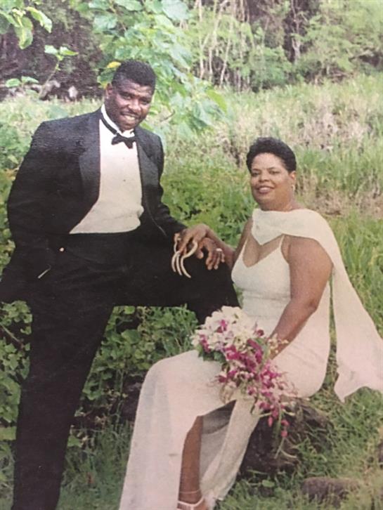 Couple wearing elegant outfits smiles joyfully in a lush garden filled with greenery.