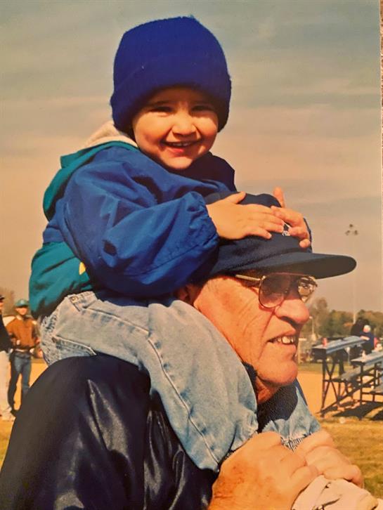 A child in a blue jacket smiles on an elderly man's shoulders, enjoying time outdoors.