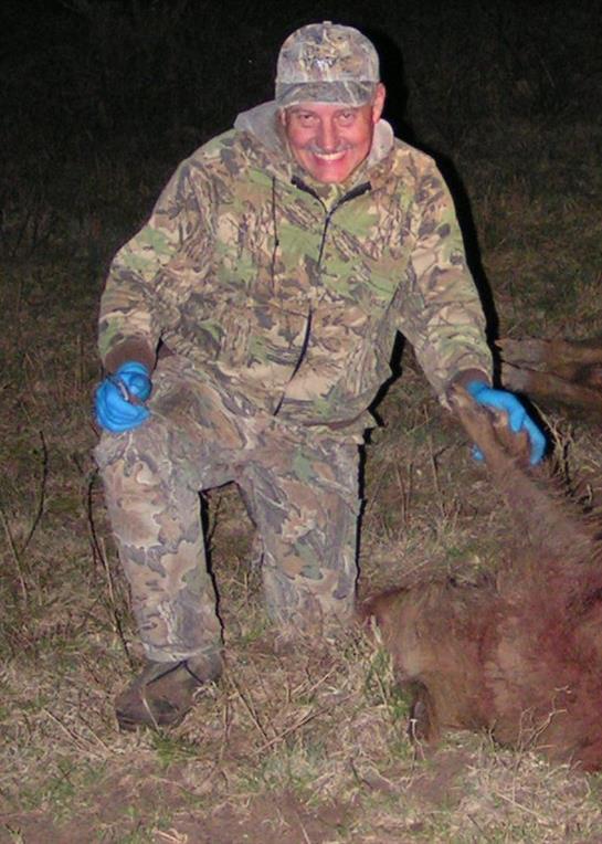 A man wearing camouflage clothing smiles while holding his hunting catch in a dark forest.