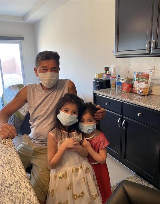 Two young girls and an adult man smile together in a bright kitchen while wearing masks.