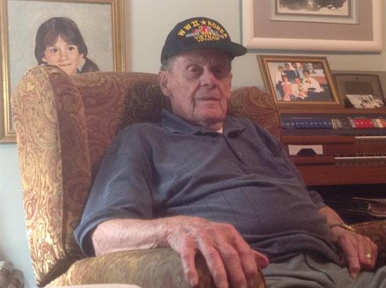 Older man with a cap sits in a cozy chair surrounded by family pictures and warm decor.