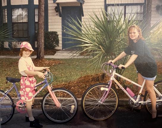 Girls are happily posing with their bicycles outside a house, enjoying a summer day.