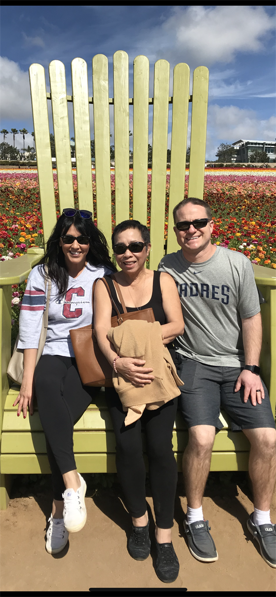Three friends sit cheerfully on a large chair surrounded by colorful flowers during daytime.