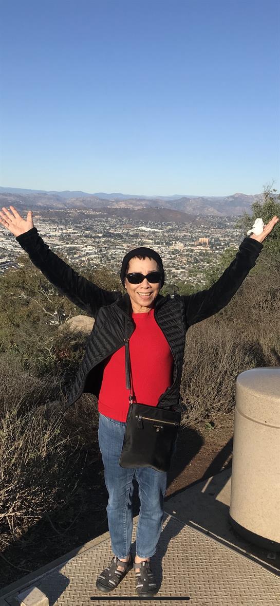 A woman in a red shirt and sunglasses stands joyfully on a hillside, arms raised, enjoying the view.
