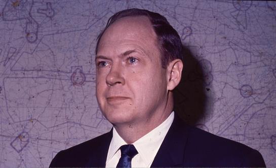 A man in a dark suit stands against a map backdrop, exuding a serious demeanor and confidence.