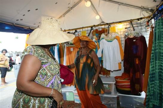 Two women in large hats browse a market stall full of colorful garments under bright lights.
