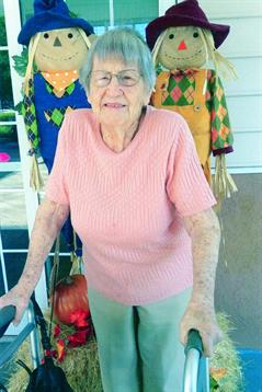 An elderly woman smiles while posing by vibrant autumn decorations on a sunny day.