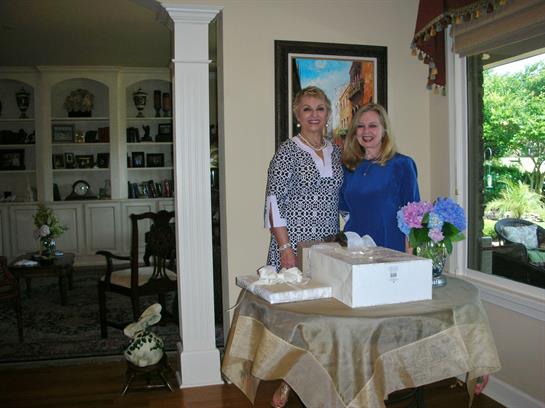 Two women stand joyfully by a table with a gift box and flowers, enjoying each other's company.