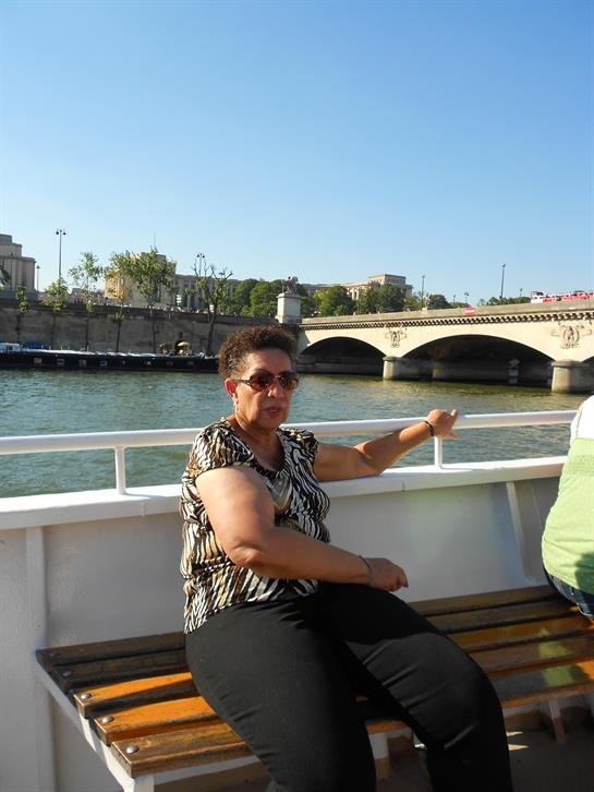 A woman relaxes on a boat, pointing toward a scenic bridge on a bright day.