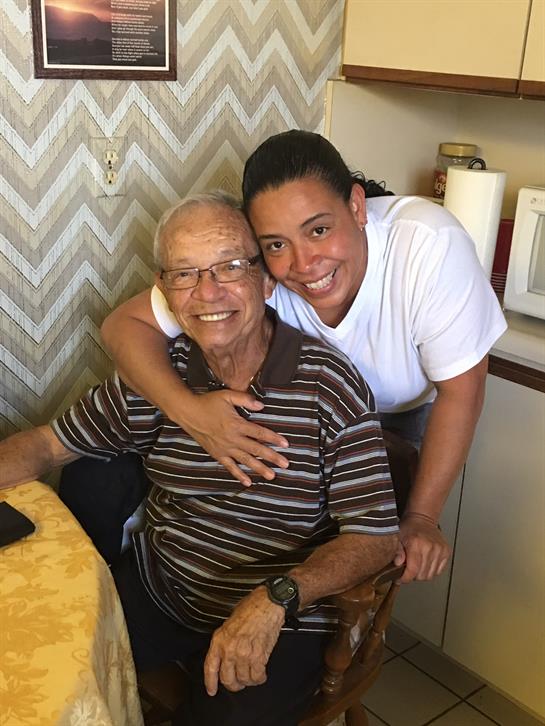 Elderly man sits happily at a table while woman stands close, embracing him with a smile.
