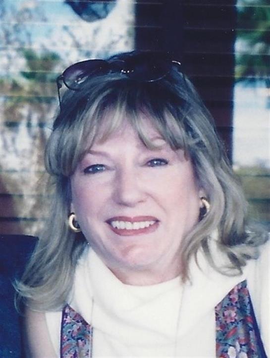 A smiling blonde woman with glasses sits indoors, radiating warmth and joy.