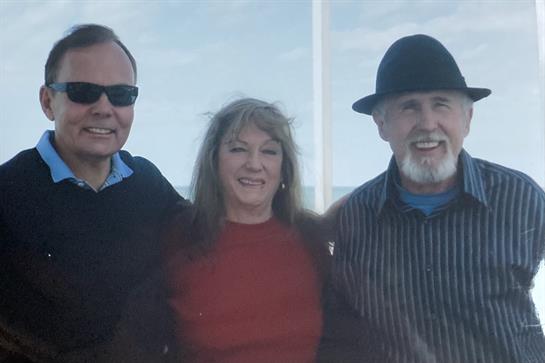 Three friends smile for a photo while enjoying a clear day by the coast, dressed casually.